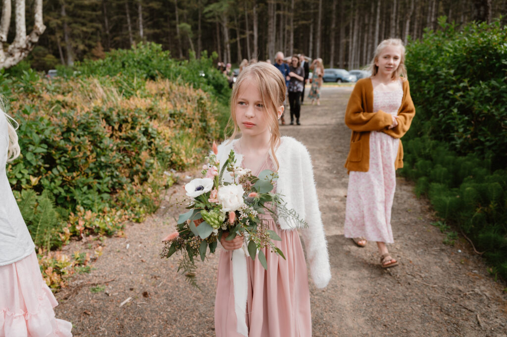 brides daughter holding her own bouquet looking pensively as she walks to the ceremony site at their intimate oregon coast wedding 